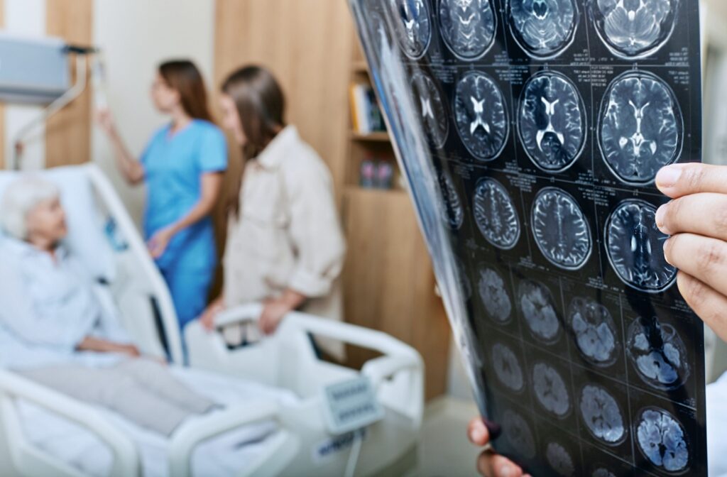 A senior in a hospital bed talks to their nurse and adult child while a doctor holds up brain scans for a dementia diagnosis